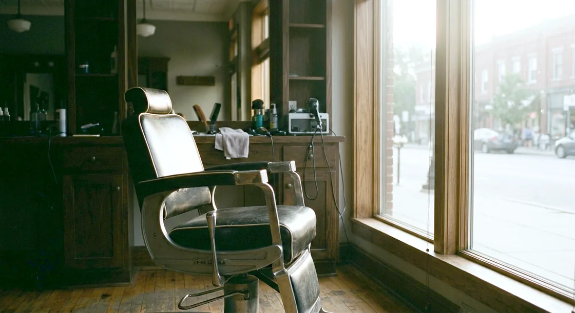 An empty barber chair beside a tall salon window in hard morning light, tools laid out on the counter behind it, the shop otherwise empty.