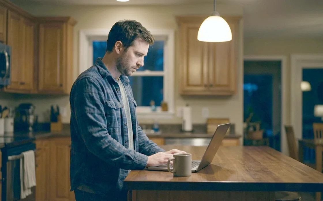 A man standing at a kitchen island late in the evening with a laptop open in front of him and a mug beside it, the house dark around him.