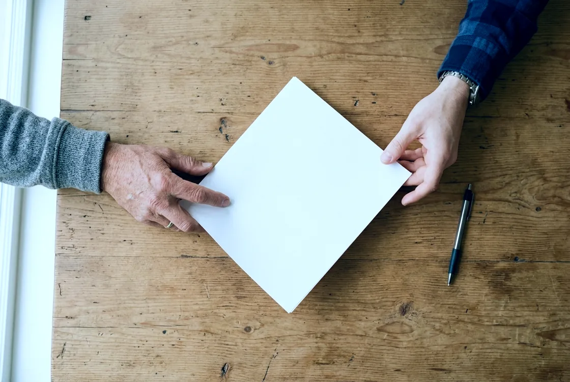 Two people&rsquo;s hands reaching in from opposite sides of a wooden table, one sliding a blank sheet of paper toward the other, photographed from directly above.